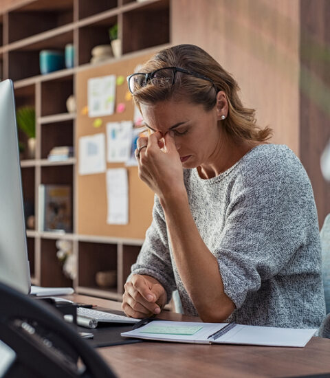 Exhausted businesswoman having a headache at office. Mature creative woman working at office desk feeling tired. Stressed casual business woman feeling eye pain while overworking on desktop computer.